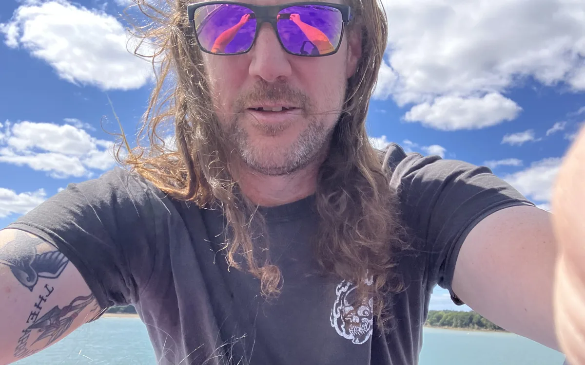 Jamie, owner and founder, by the sea: long hair, sunglasses, tattooed arms.